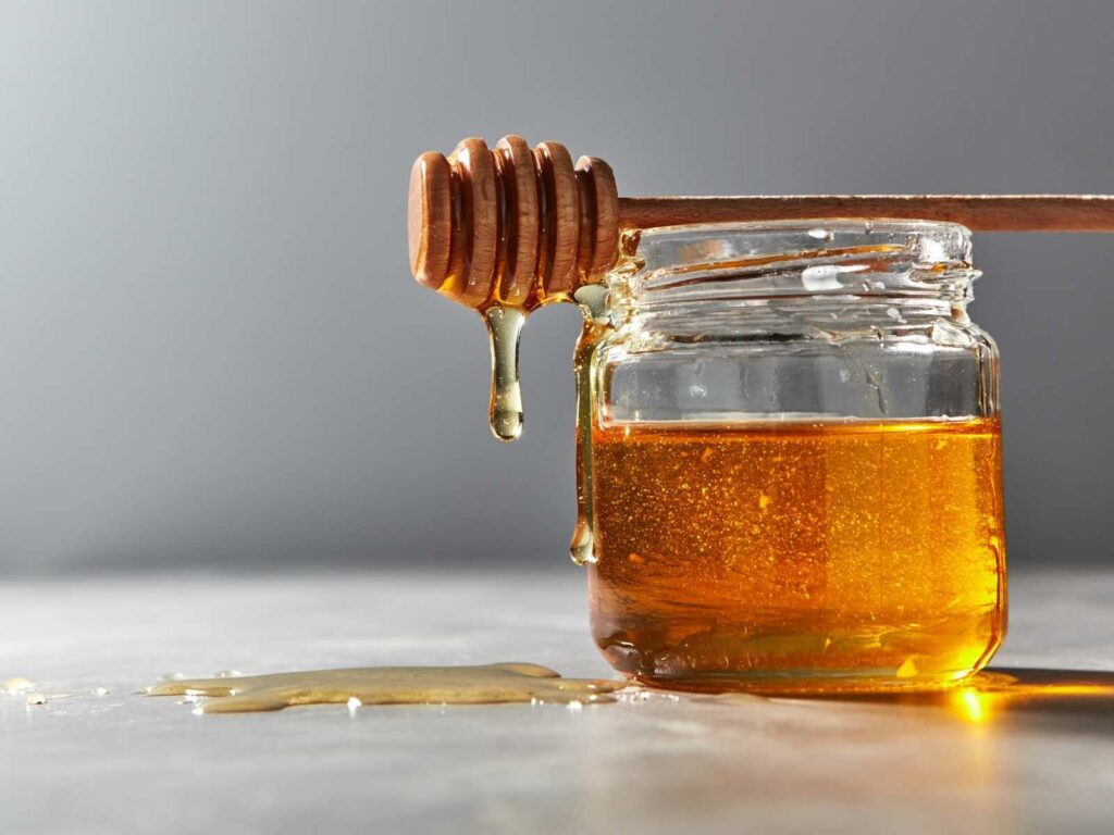wide glass jar with honey on a table with wooden honeycomb resting on the top with honey dripping off onto table
