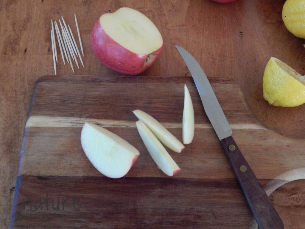wooden cutting board on a wooden table with a sharp knife next to four apple slices half of an apple on the table with toothpicks and a lemon cut in half in the corner