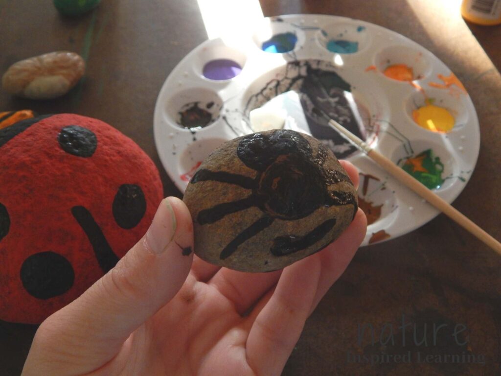 hand holding a bee painted rock with black paint on the rock ladybug painted rock on table with moth painted rock paint tray with a variety of acrylic paints on the tray with a paint brush with black paint resting on tray yellow acrylic paint bottle in top corner