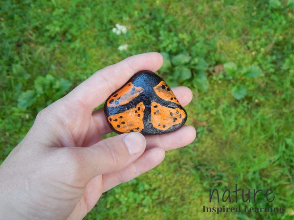 hand holding a DIY handmade rock art monarch butterfly painted rock outside with green background