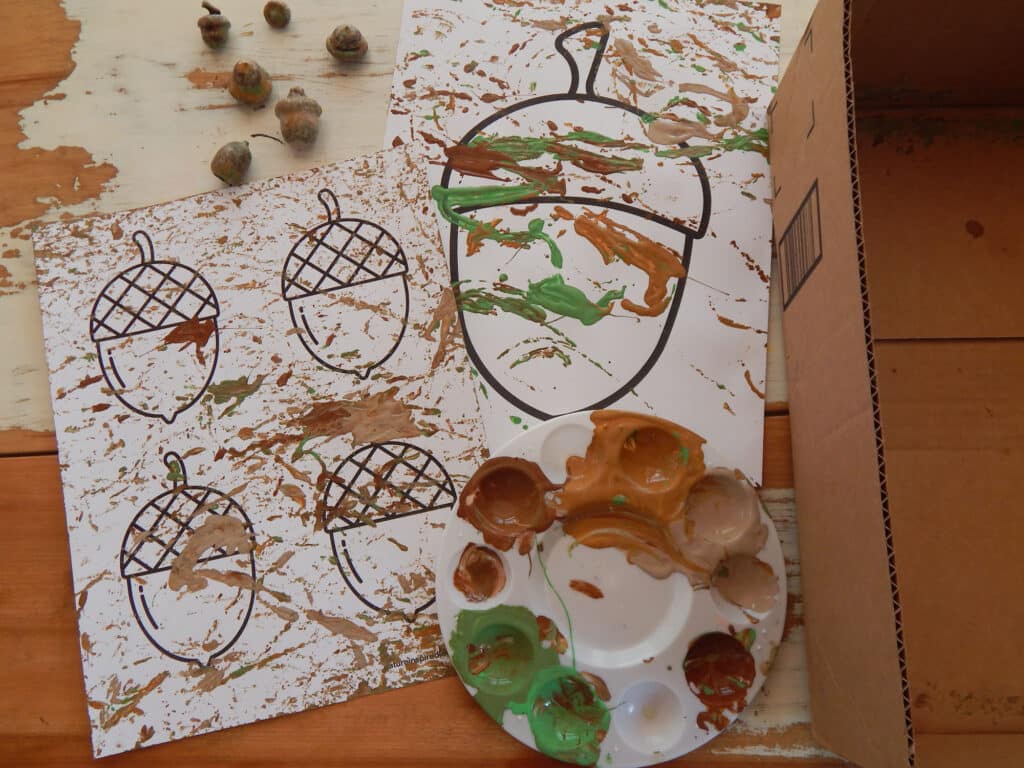 two acorn coloring pages on a table covered in paint with a paint tray with paint and six real acorns on the table along with a cut cardboard box