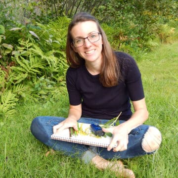 Woman outside siting in the grass wearing jeans and a navy blue shorts sleeve black rimmed glasses hair down holding a nature journal with coloring pencils, greenery, field microscope ferns and foliage in background