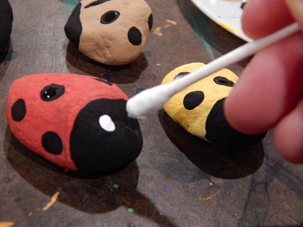 hand holding a q tip with white paint on the end applying eyes to handmade red ladybug painted stone tan and yellow ladybug on the table without eyes