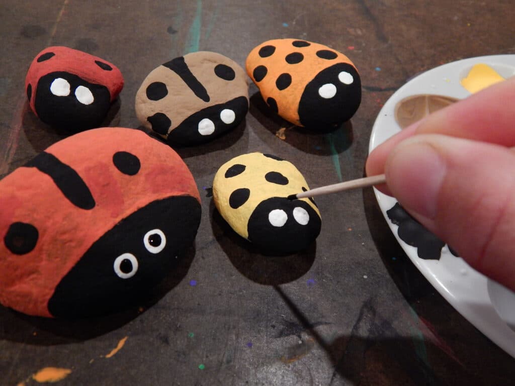 hand holding a toothpick with black paint on the end adding eyes to handmade yellow lady beetle painted stone with four more lady beetle stones next ton the table paint tray to the side