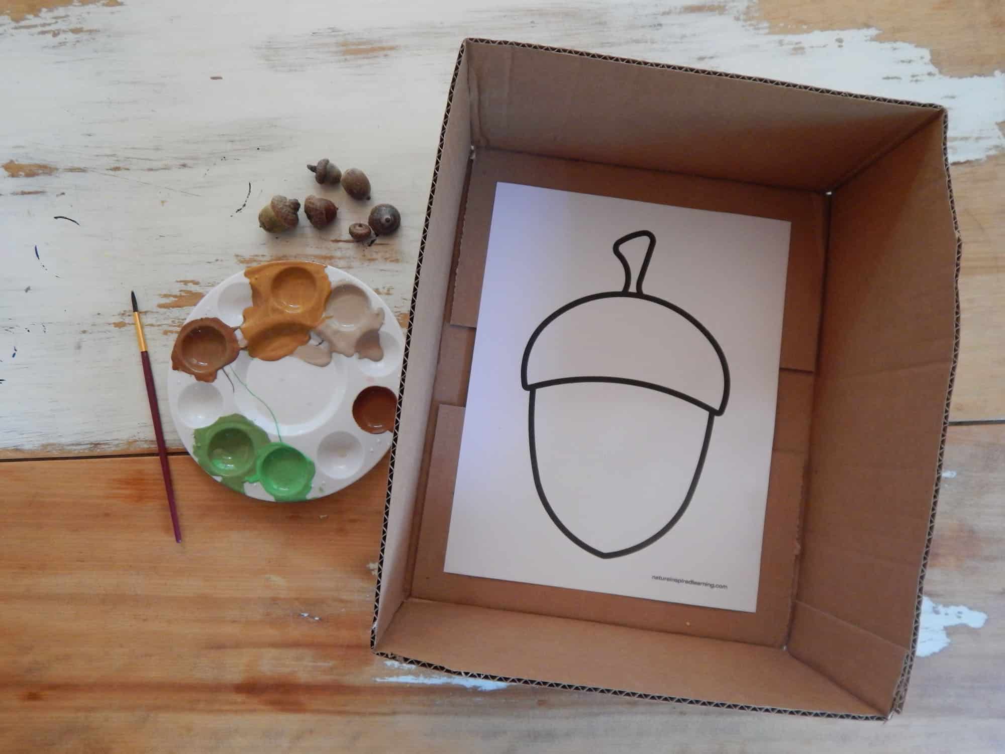 a cut cardboard box with an acorn template inside on a wooden table with a plastic paint tray with orange, tan, brown, and green paint a paint brush, and six real acorns