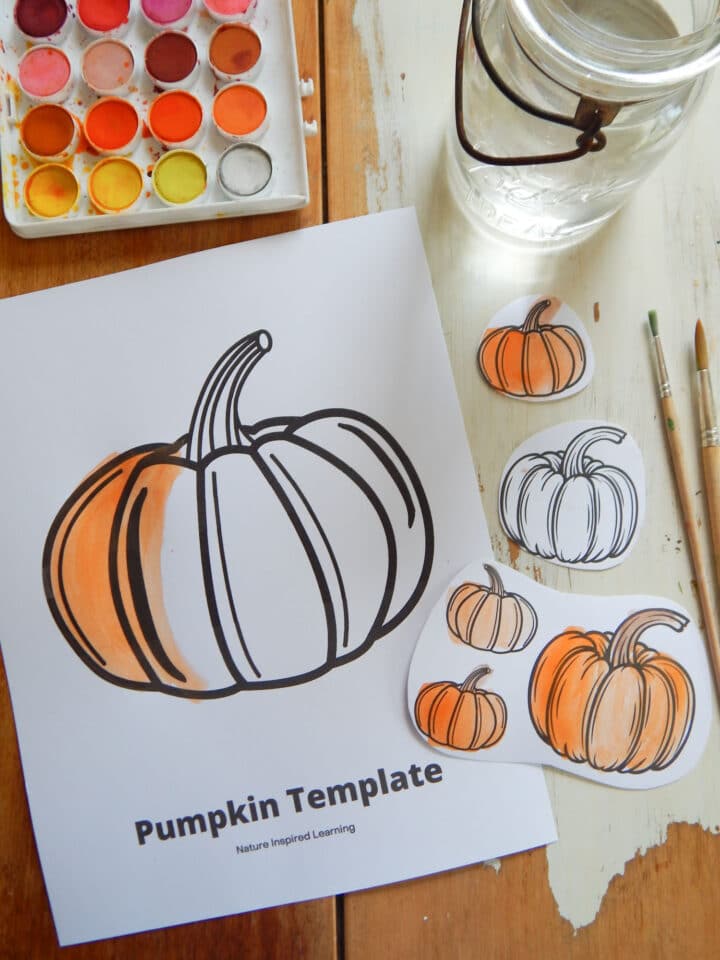 pumpkin coloring page on a wooden table partially colored in orange with watercolor paint set, water, two paint brushes, and five little cut out paper pumpkins