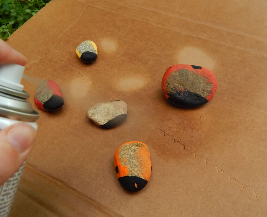 hand spraying clean protective sealer on hand painted beetle rocks turned upside down to seal the bottom five rocks on cardboard outside