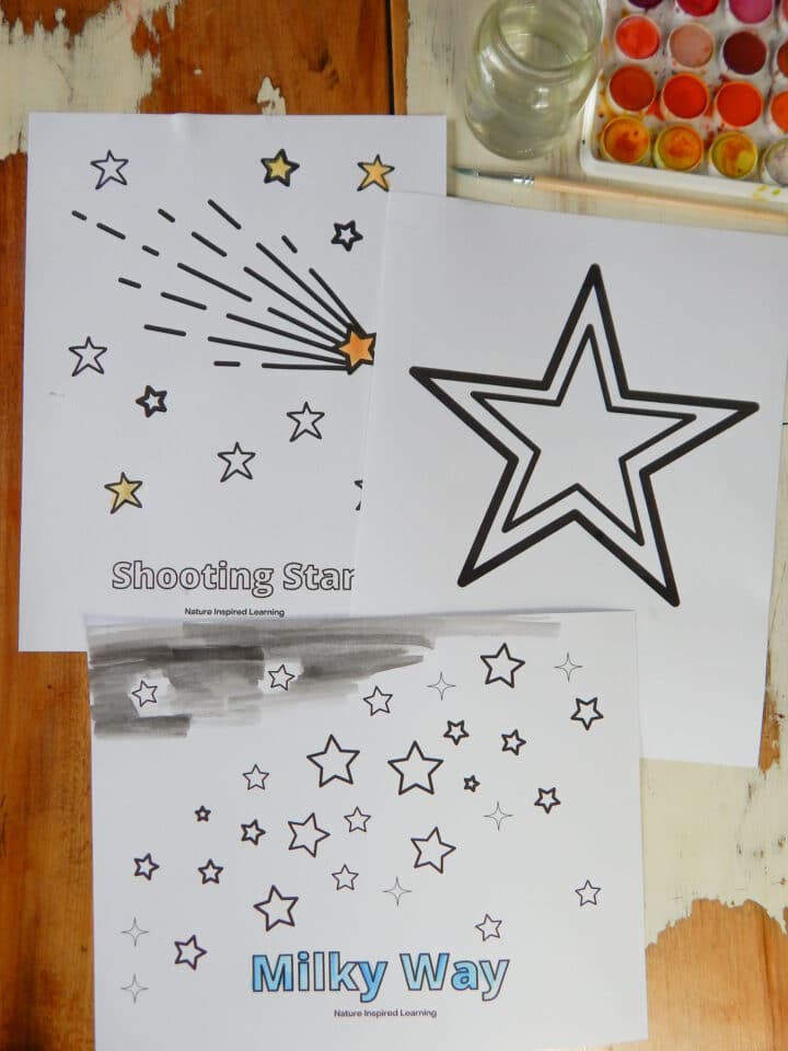 collection of star coloring pages printed out on a wooden table partially colored in using watercolor paint. Large star, milky way, and shooting star sheets. Glass jar with water above, wooden paint brush, and watercolor set top corner