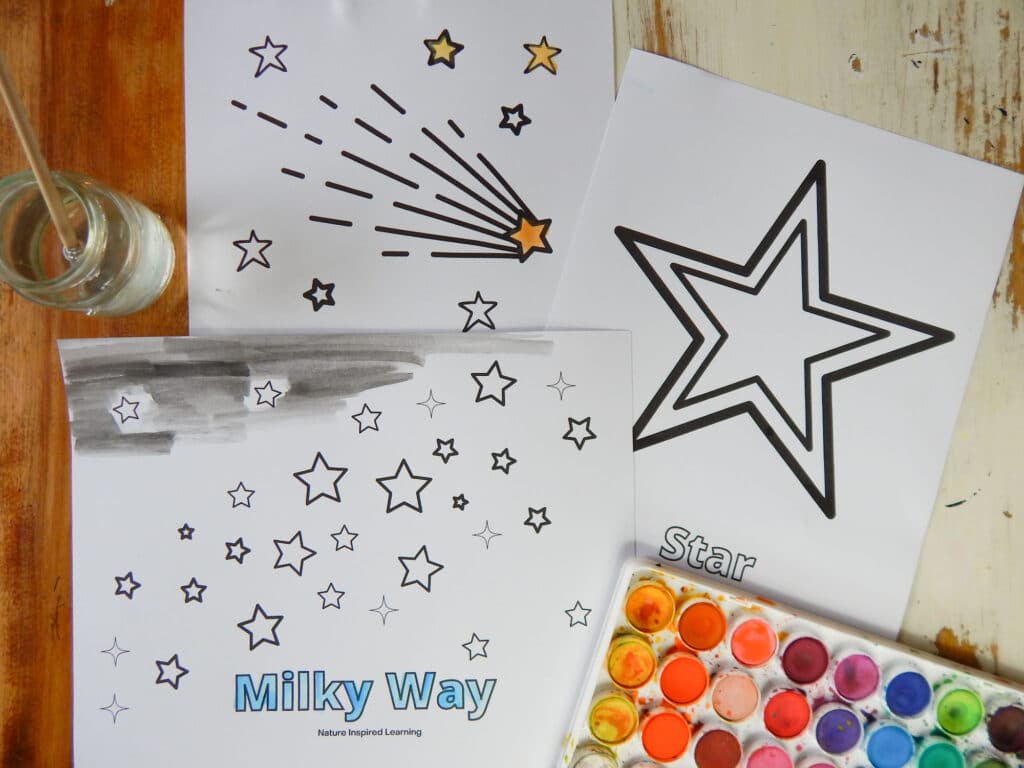 Shooting star coloring page, large star coloring page, and milky way with collection of stars all overlapping on wooden table small glass jar with wooden paint brush in it the the left and bottom right watercolor paint set. Coloring pages partially painted in