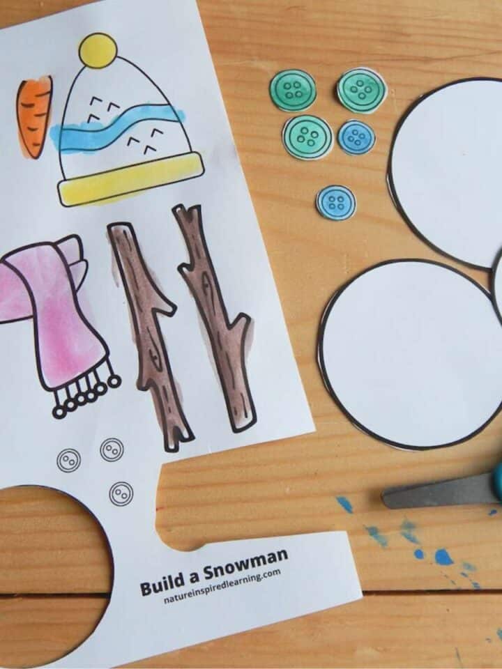 build a snowman printable colored in and half cut out on a wooden table. Cut out snowballs and buttons with kid scissors to the side.