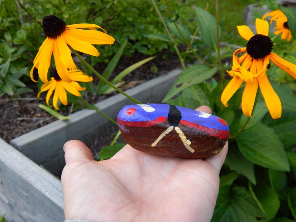 hand holding a hand painted butterfly painted rock to show the wings and antenna outside in the garden with blooming blackeyed susans and green plants