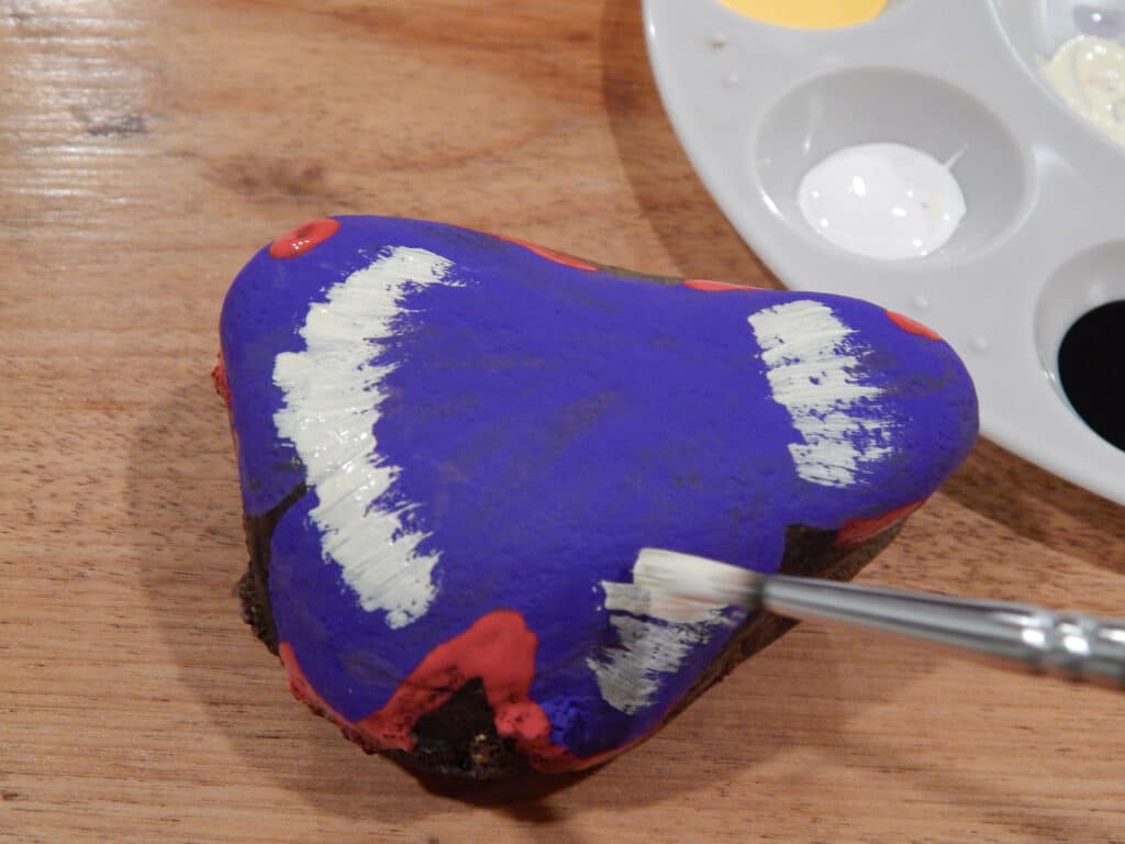 paint brush with white paint against a butterfly painted rock being used to add a pattern to the wings. Paint tray with paint on wooden table