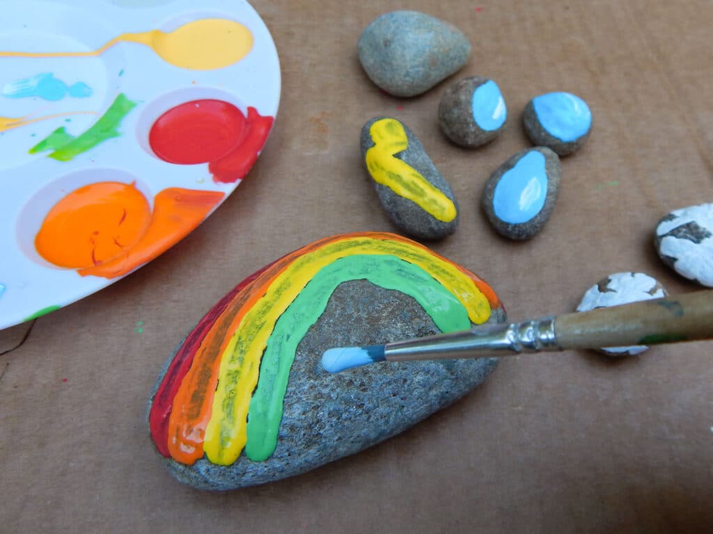 paint brush with blue paint above a rainbow design being painted on a rock. Paint tray with orange, red, yellow, green, and blue paint. One unpainted rock with six small painted rocks, lightning bolt, water drops, and snowflakes. All on cardboard.