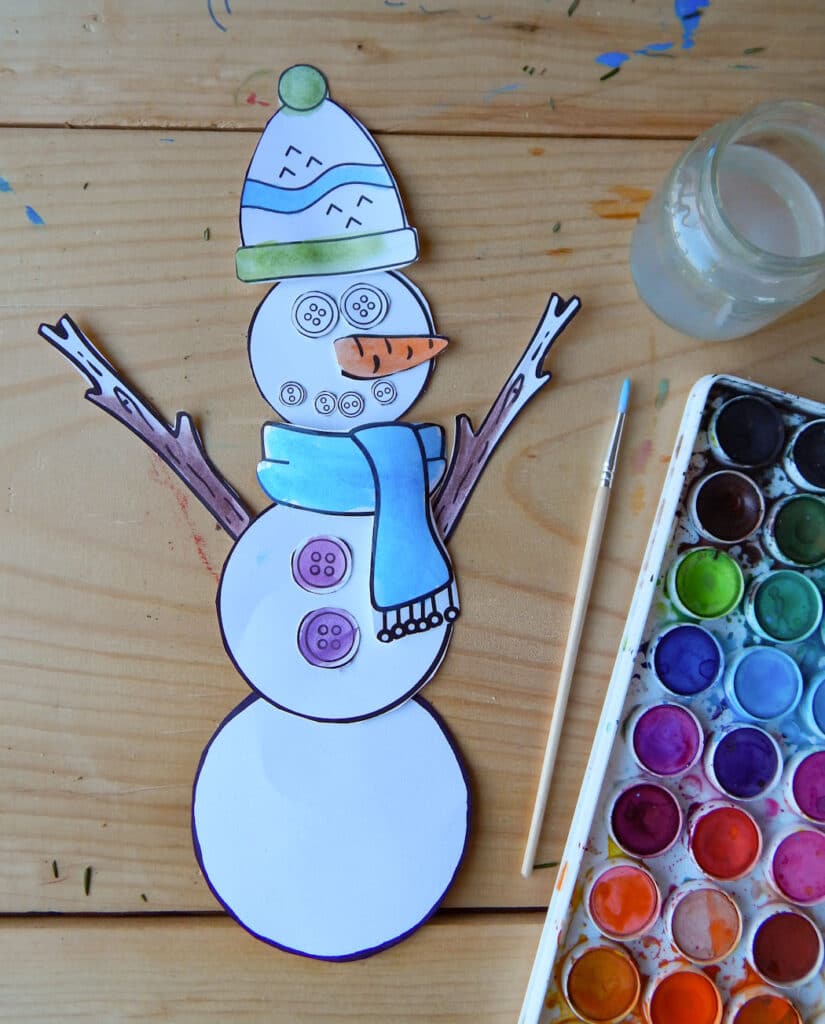 paper snowman craft cut out and built on a wooden table. Watercolor paint set, glass jar, and paint brush to the side. Color added to the printable using watercolor paint.