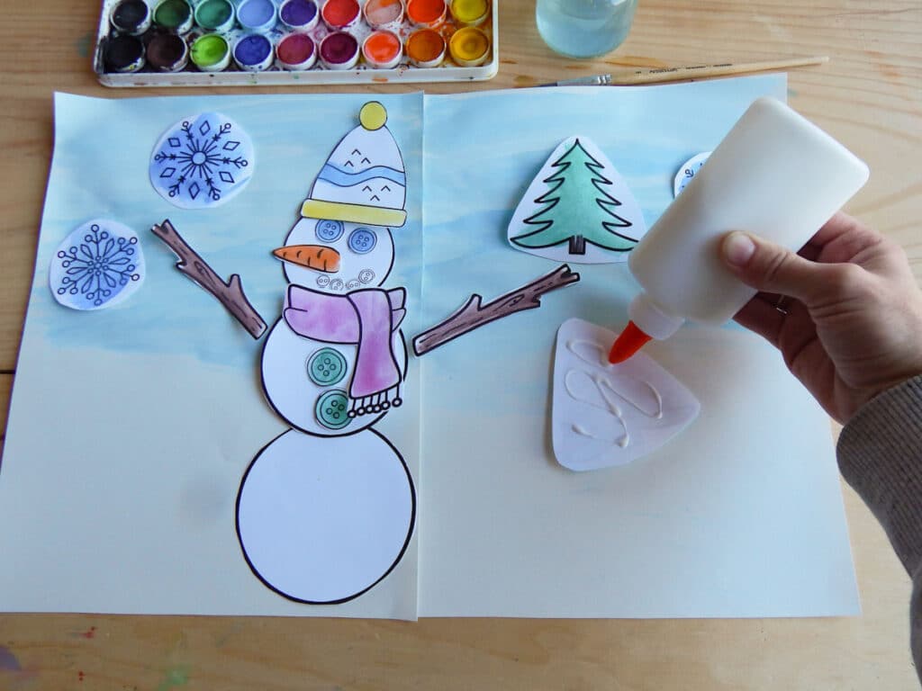 hand applying glue to the back of paper for a paper snowman craft. Watercolor paint set with jar of water above paper with snowman scene