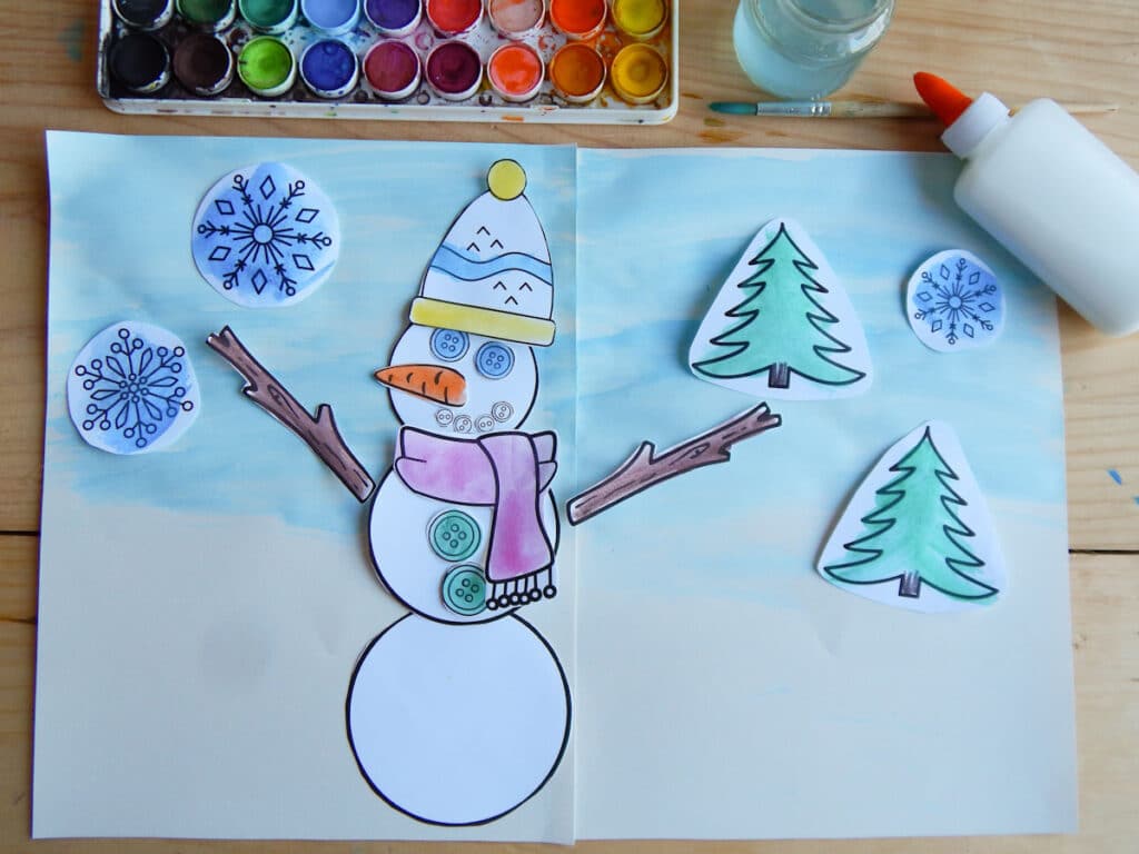 completed build a snowman paper craft for kids on a wooden table with watercolor paint set, glass jar with water, paint brush, and school glue. Paper snowman glued down to winter scene with paper snowflakes and evergreen trees