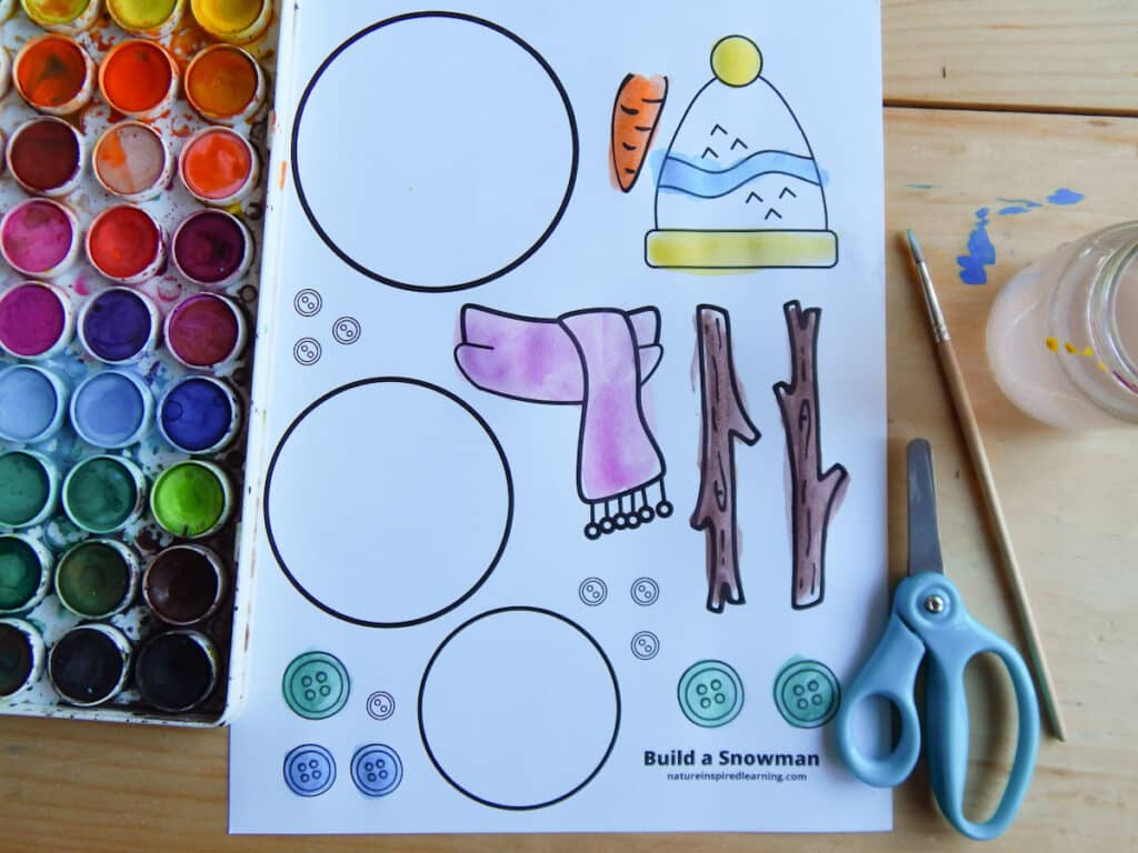 build a snowman printable on a table with watercoloring paint set, scissors, paint brush, and glass jar with water. Printable colored in using paint