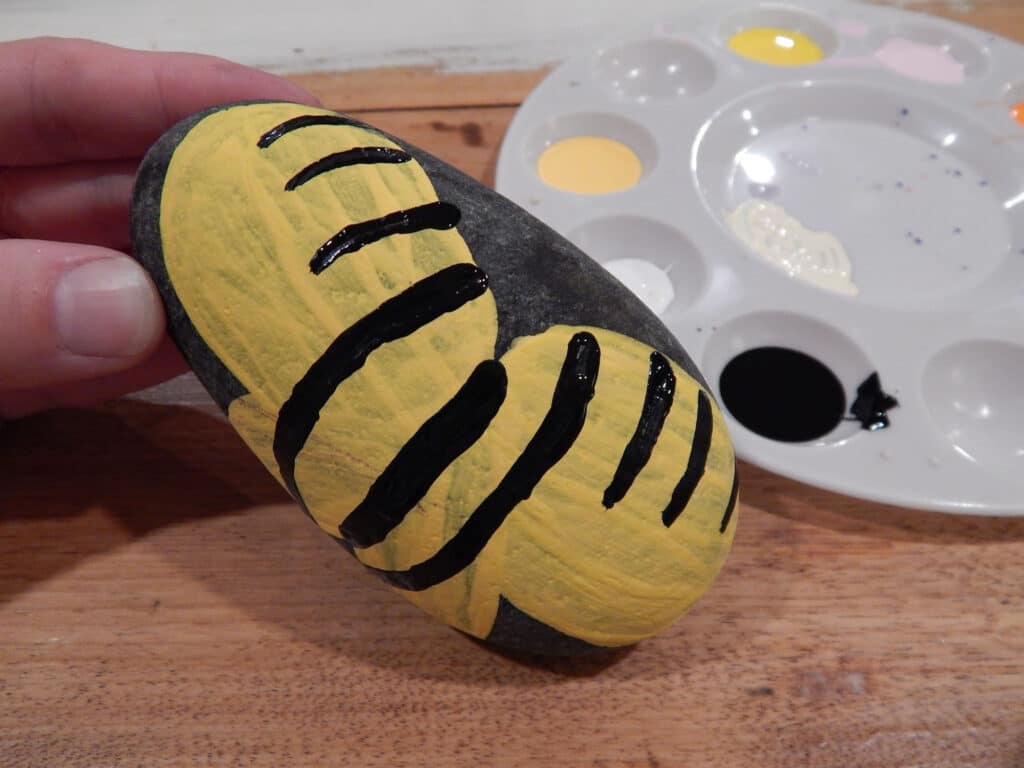 hand tilting up a butterfly painted rock after painting black stripes on a yellow body. Paint tray with paint in background on table