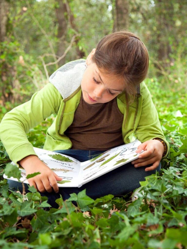 child sitting outside on the ground reading a book about plants surrounded by green plants