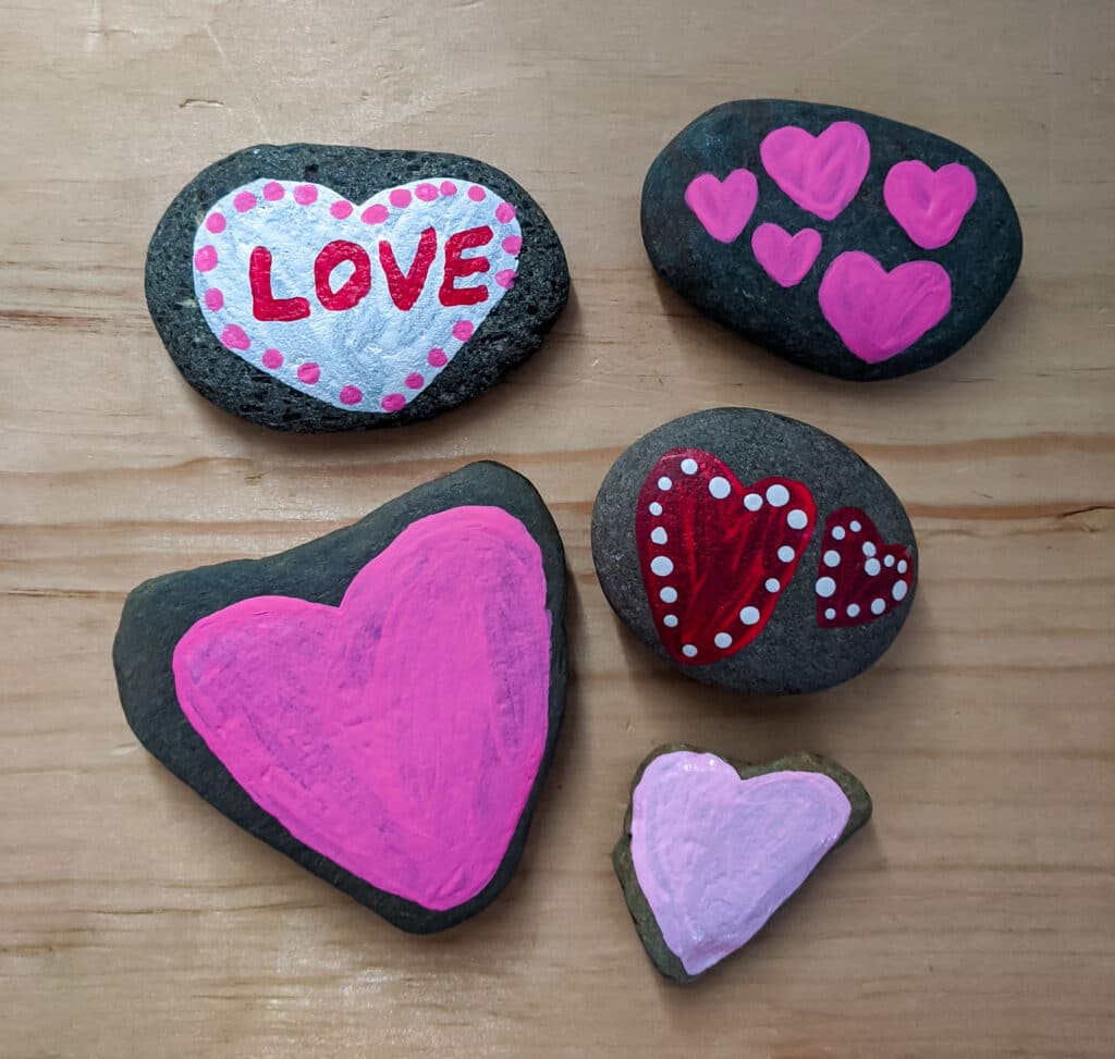 handmade heart painted rocks with different designs on a wooden table. Light pink heart, bright pink heart, red hearts with white dots, mini pink hearts, and white heart with red love and pink dots