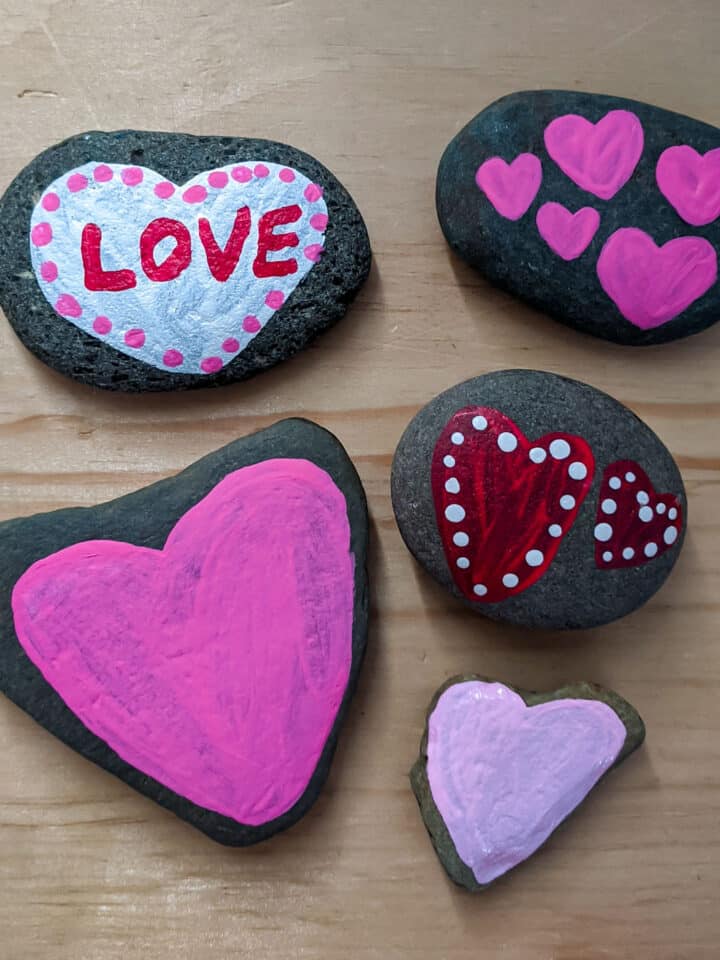 handmade heart painted rocks with different designs on a wooden table. Light pink heart, bright pink heart, red hearts with white dots, mini pink hearts, and white heart with red love and pink dots
