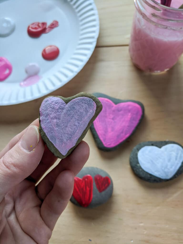 hand holding a heart shaped rock painted light pink. Below on the wooden table are three rocks painted with heart designs, paint on a paper plate and a glass jar with pink water and paint brushes in it