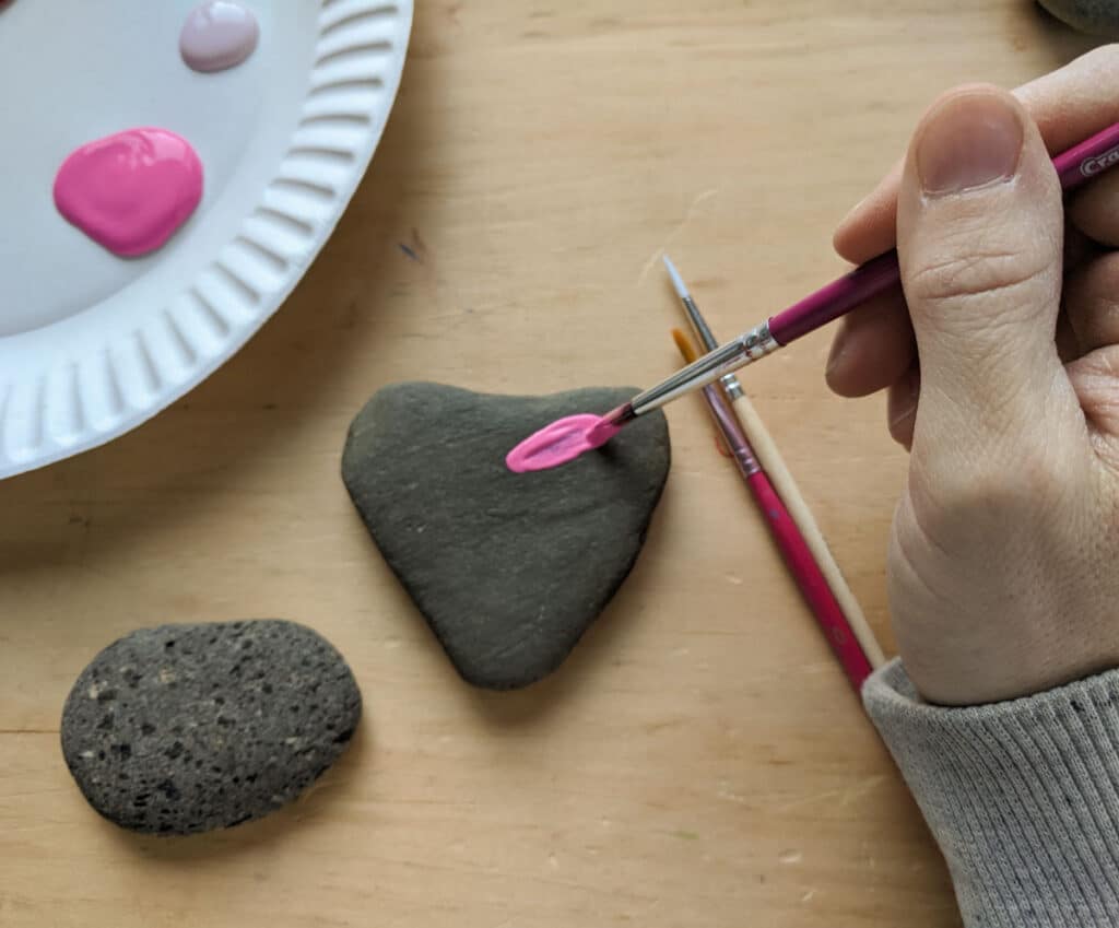 hand holding a paint brush applying pink paint on a heart shaped rock. Painting supplies and another rock on the table below