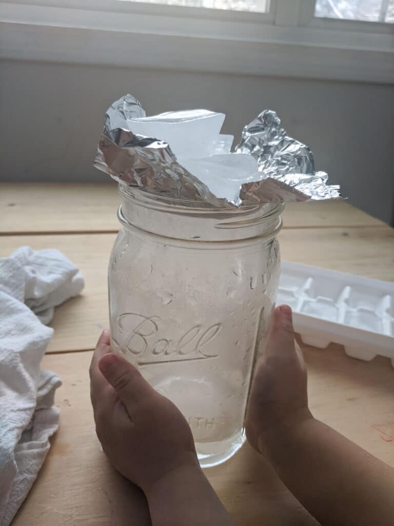 child's two hands wrapped around a wide mouth glass jar with water inside and fog. Foil lid on top of jar with collection of ice cubes. Ice cube tray and white towel on wooden table on either side of the jar.