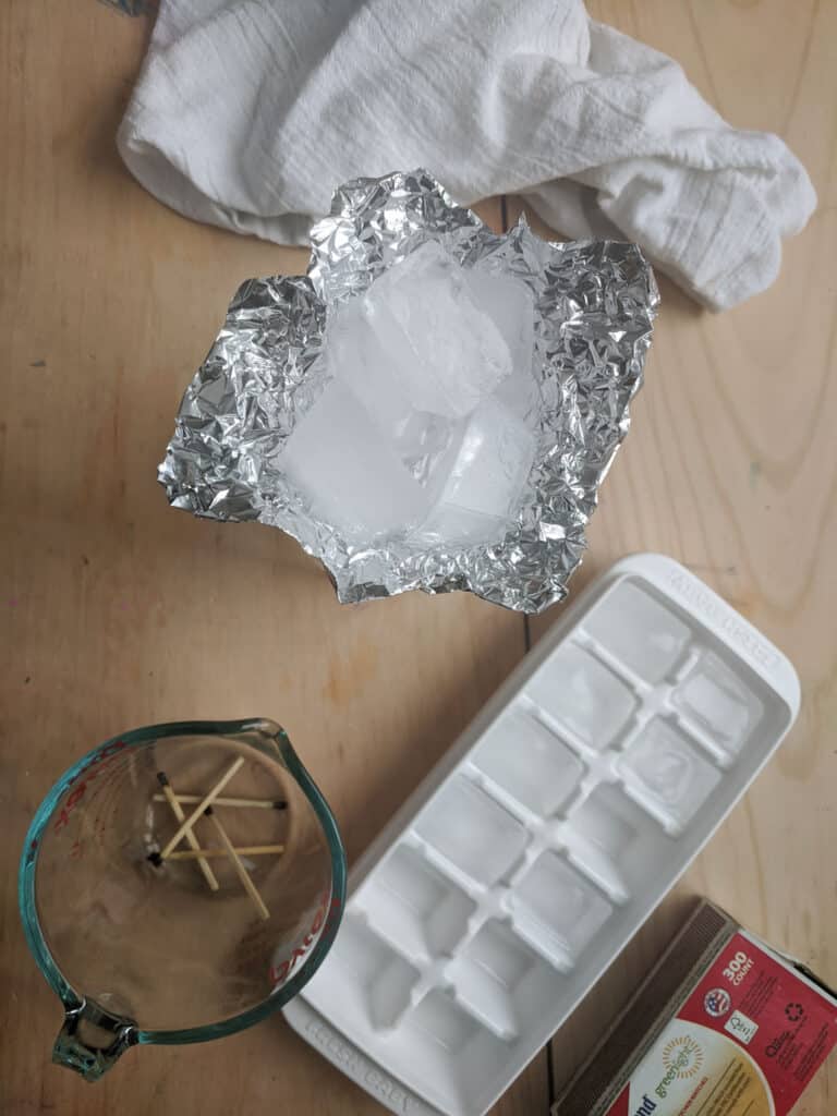 supplies on a wooden table including white towel, tin foil in a cup shape holding ice cubes, ice cube tray, glass measuring cup with used matches, and a box of matches