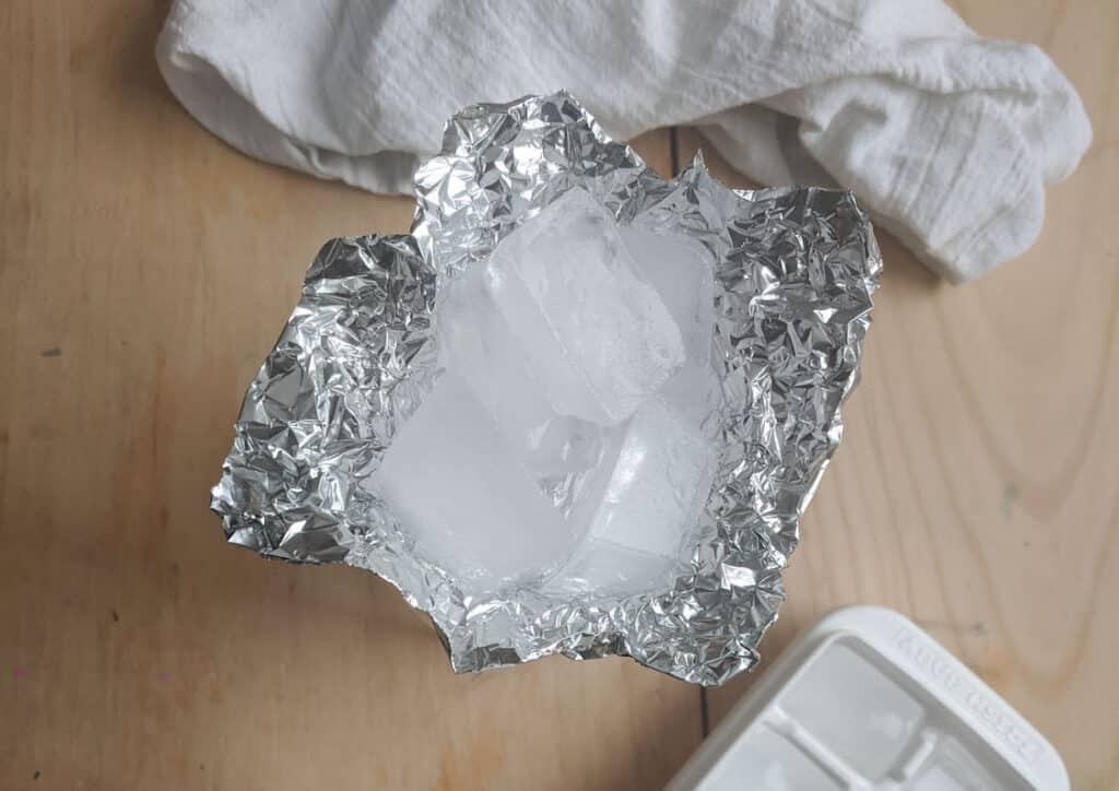 aluminum foil shaped into a bowl with four ice cubes inside. White towel on table above and white ice cube tray with ice cubes below.