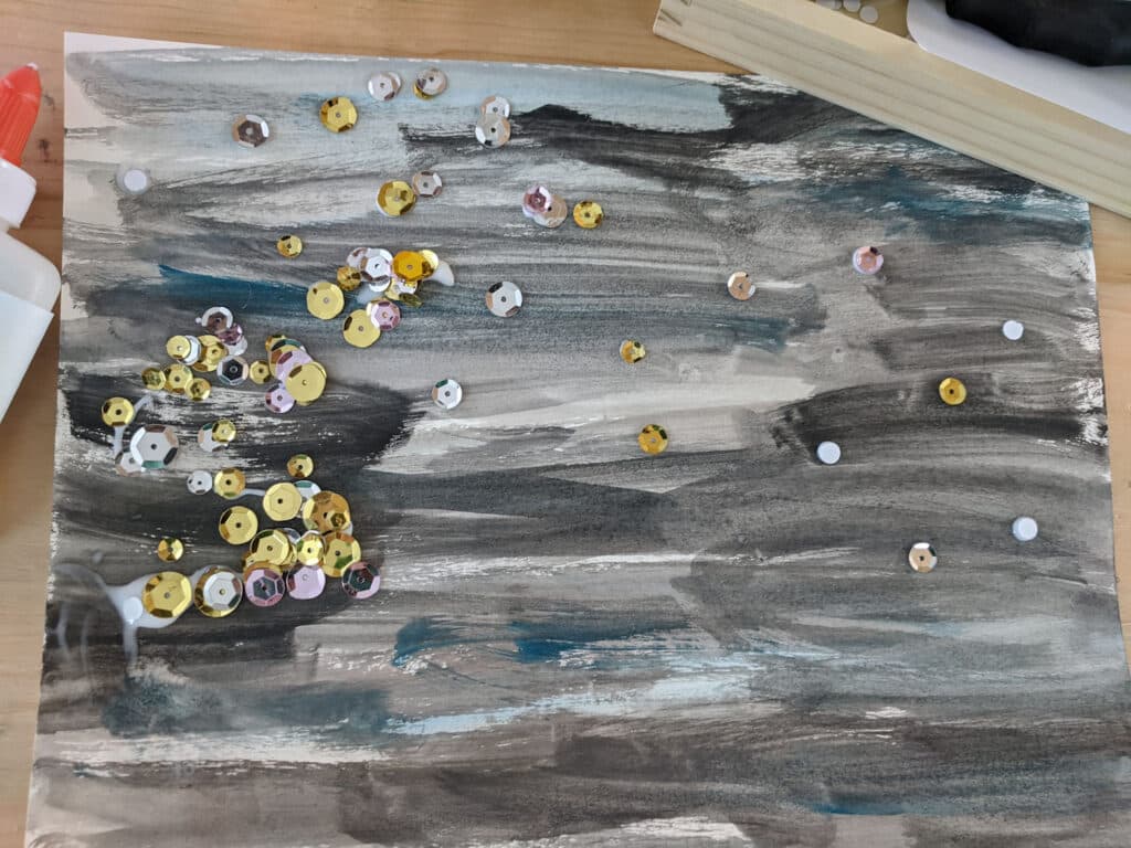 a night sky watercolor process art with different gold, silver, and pink sequins glued down on a wooden table. Liquid school glue left side, wooden box top right.