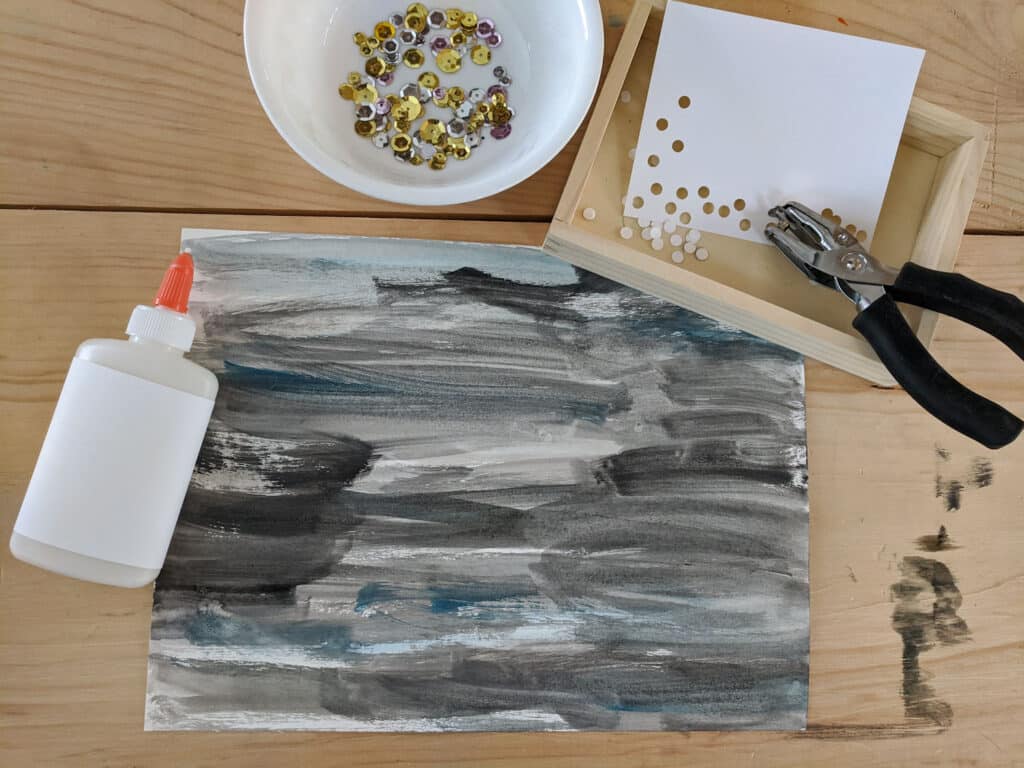 black, grey, and blue watercolor paint lines covering a piece of white paper with liquid school glue left side, bowl with gold and silver sequins above, wooden box with paper punched with hand held hole punch. All on a wooden table.