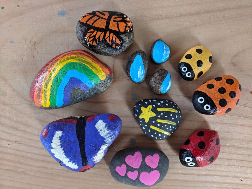 collection of painted rocks on a wooden table including monarch butterfly, raindrops, rainbow, three different colored ladybugs, mini hearts, shooting star, and a purple butterfly