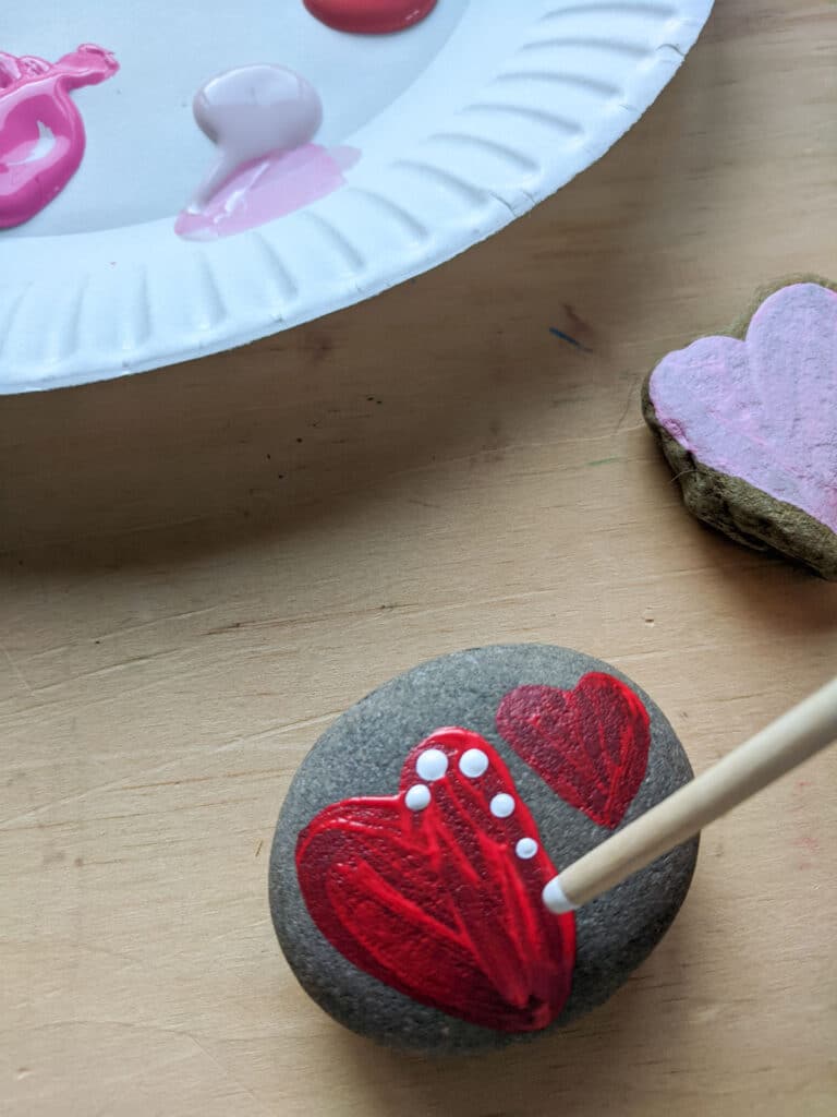 end of a paint brush with white paint on it adding dots to a red heart painted on a stone. Pink, light pink, and red paint on paper plate above with another heart shaped rock to the side