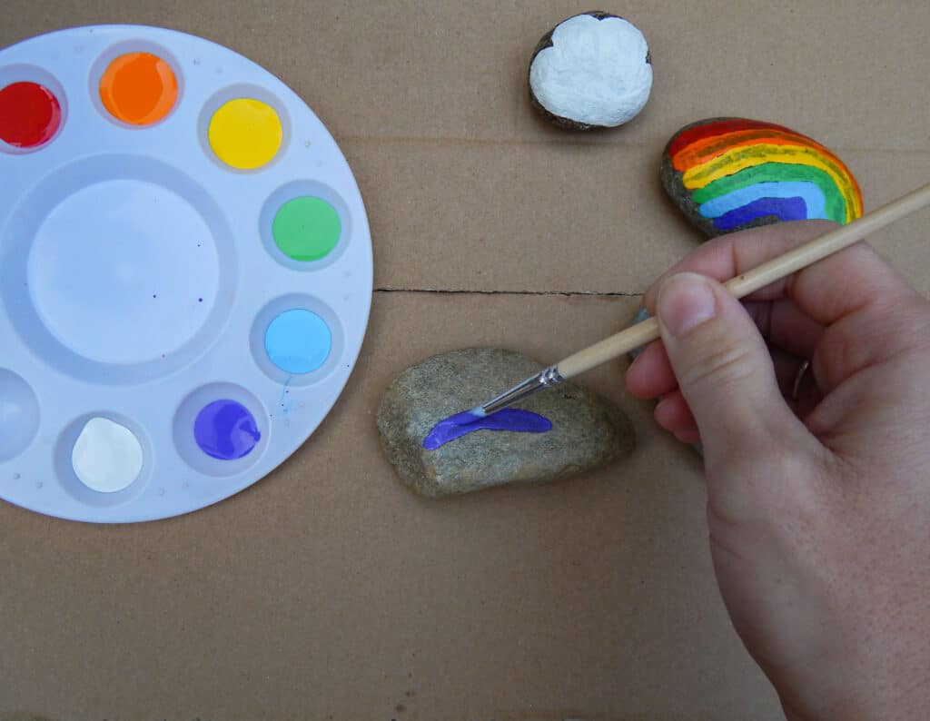 hand using a small paint brush to paint a purple band on a rock. Finished rainbow rock top right and a white cloud painted on a rock. Circular paint ray with red, orange, yellow, green, blue, purple, and white paint in each section. All supplies on cardboard.