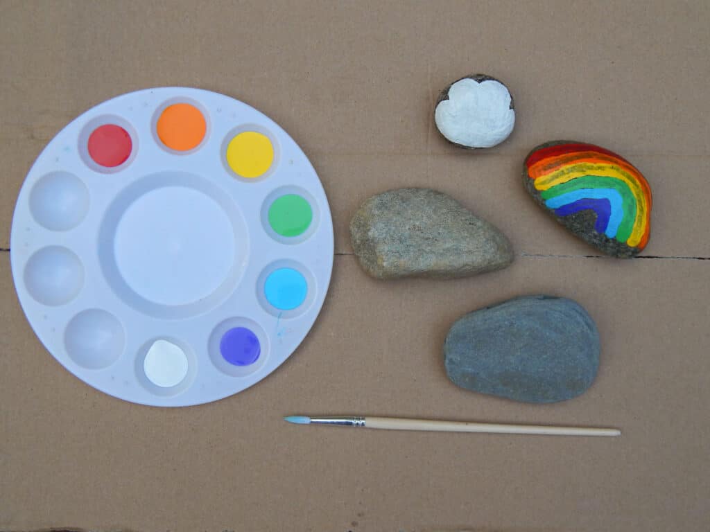 Rainbow rock painting supplies on cardboard. Circular paint tray with each color of the rainbow ROYGBIV acrylic paint next to two smooth unpainted rocks, a white cloud painted on a rock, and an arched rainbow rock painting. Small wooden paint brush below.