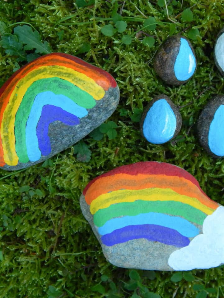 painted weather rocks outside one rainbow painted in an arch, half a rainbow with a white cloud at the end, three raindrop painted rocks, and one white cloud painted on a stone. All outside in the green grass.