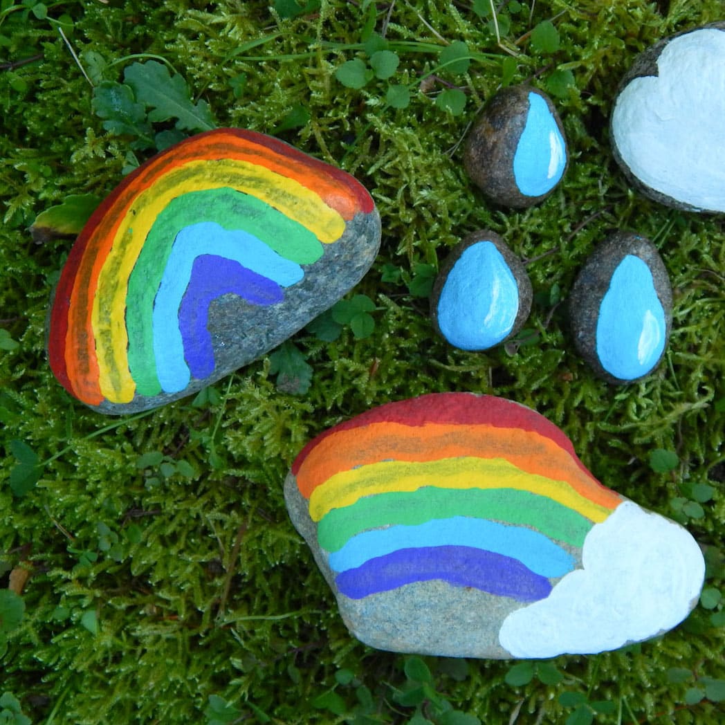 painted weather rocks outside one rainbow painted in an arch, half a rainbow with a white cloud at the end, three raindrop painted rocks, and one white cloud painted on a stone. All outside in the green grass.