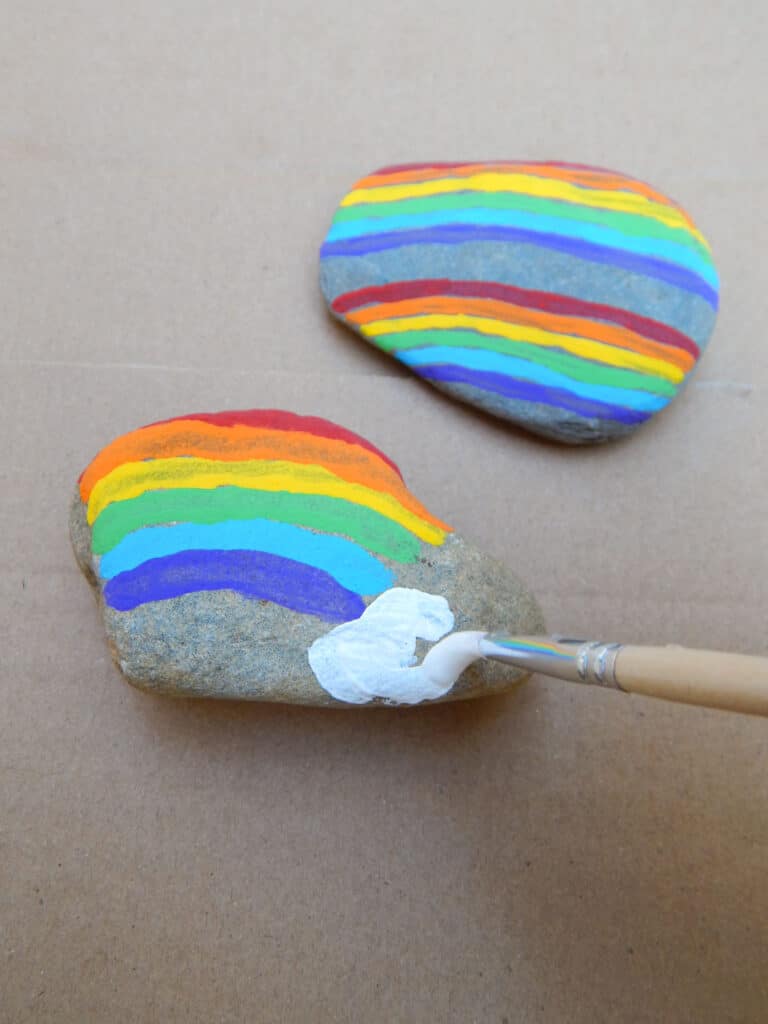 hand holding a small paint brush making a white cloud at the end of a rainbow painted on a rock. Double rainbow painted rock above. Both rocks on cardboard.