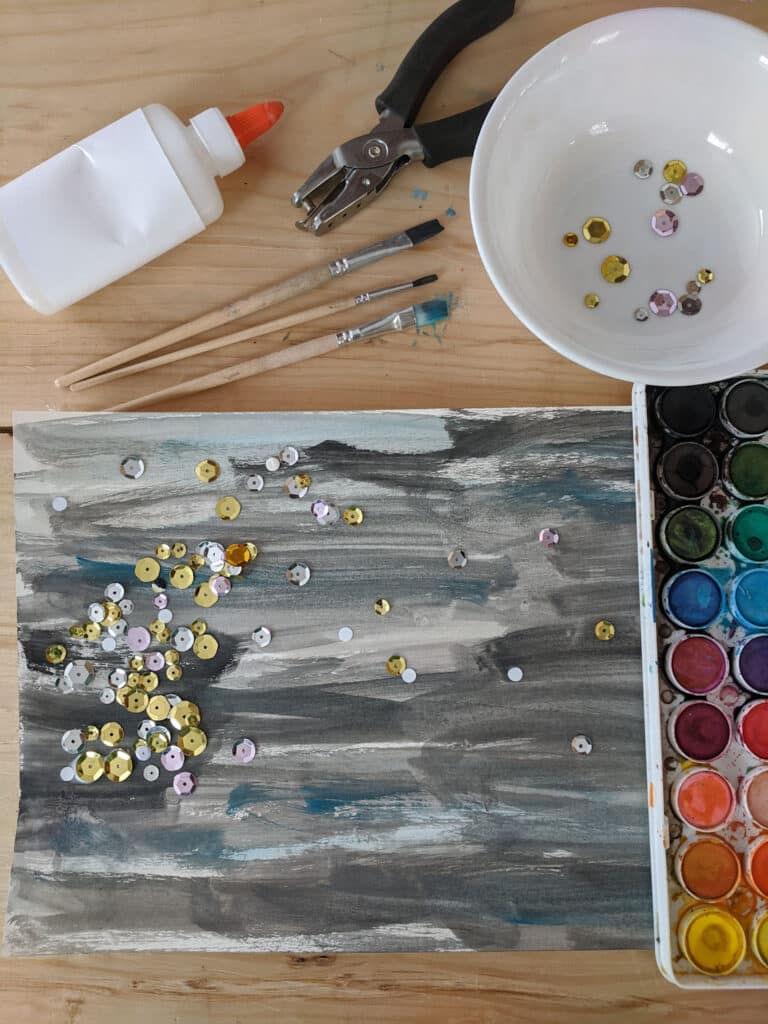 starry night process art on wooden table with supplies: liquid school glue left, three wooden paint brushes top, hand held hole punch, white bowl with sequins, and watercolor paint set right
