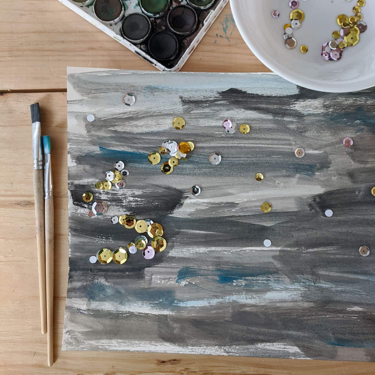 starry night process art on a wooden table made with watercolor paint, sequins, and glue. Two wooden paint brushes right side, watercolor paints above, white bowl with sequins top right.