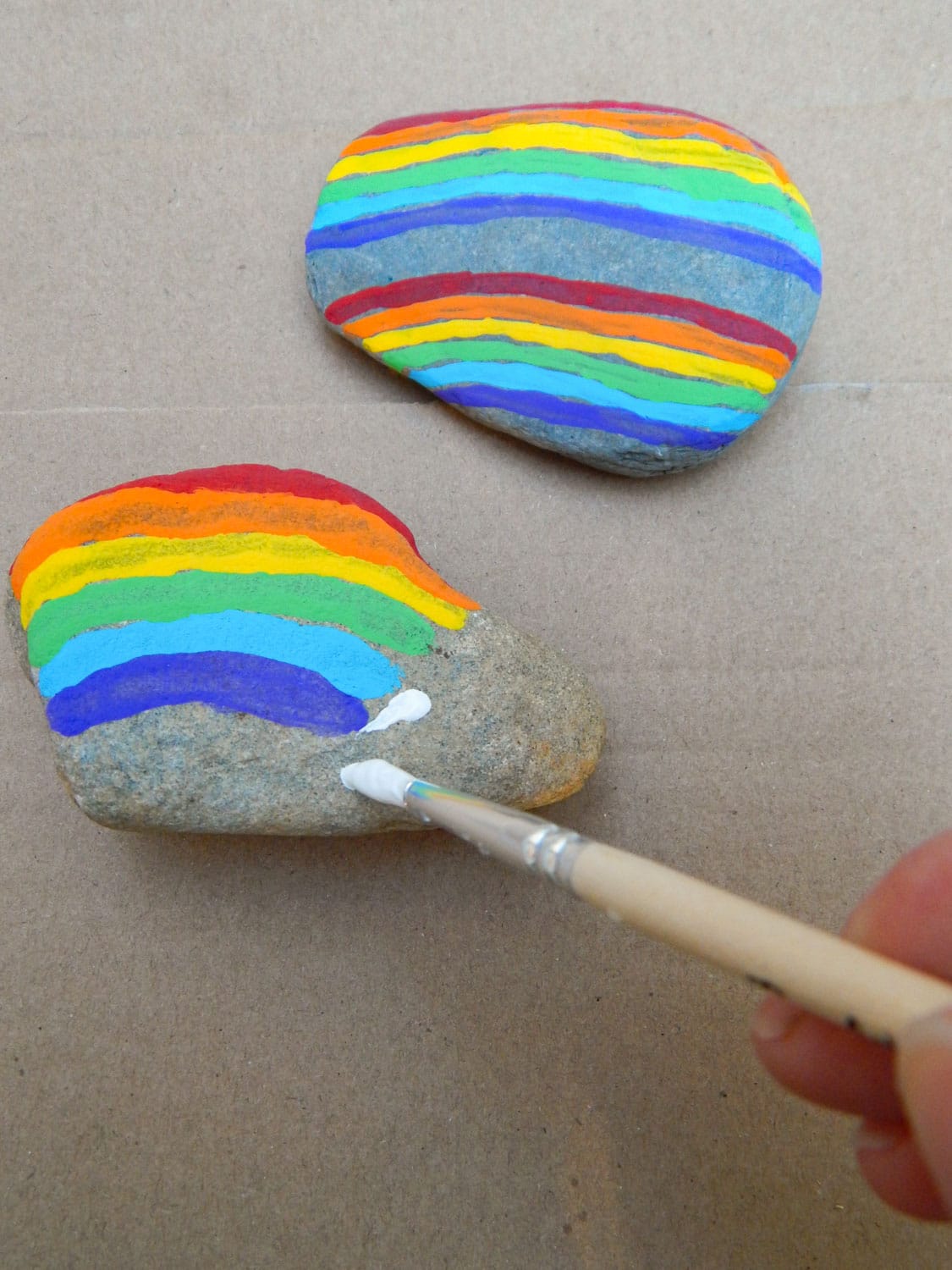hand holding a small paint brush making a white cloud at the end of a rainbow painted on a rock. Double rainbow painted rock above. Both rocks on cardboard.