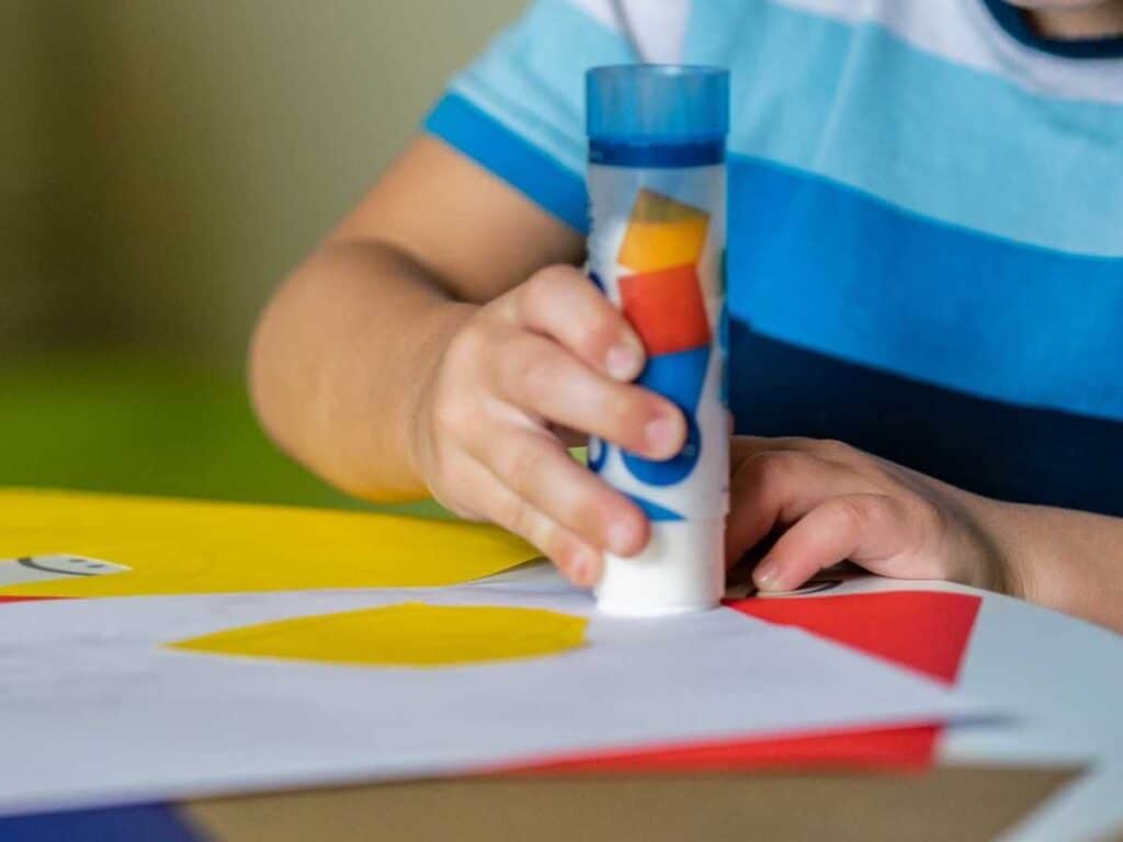 child wearing blue stripped shirt using a glue stick to apply glue to paper cut at the table