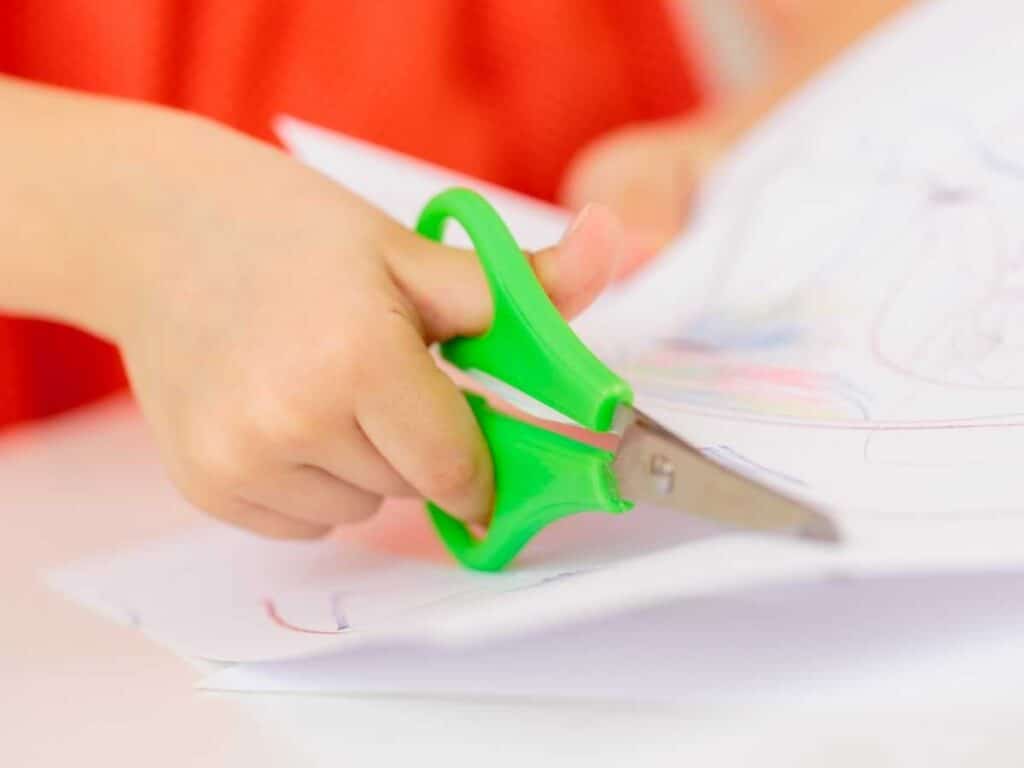 young child wearing orange shirt cutting paper with green safety scissors at a table