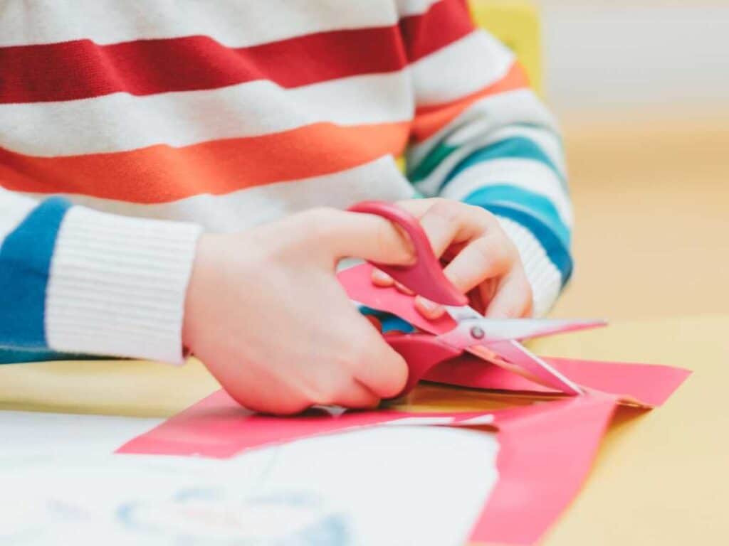 child wearing a rainbow striped sweater cutting red paper with pink scissors at a desk