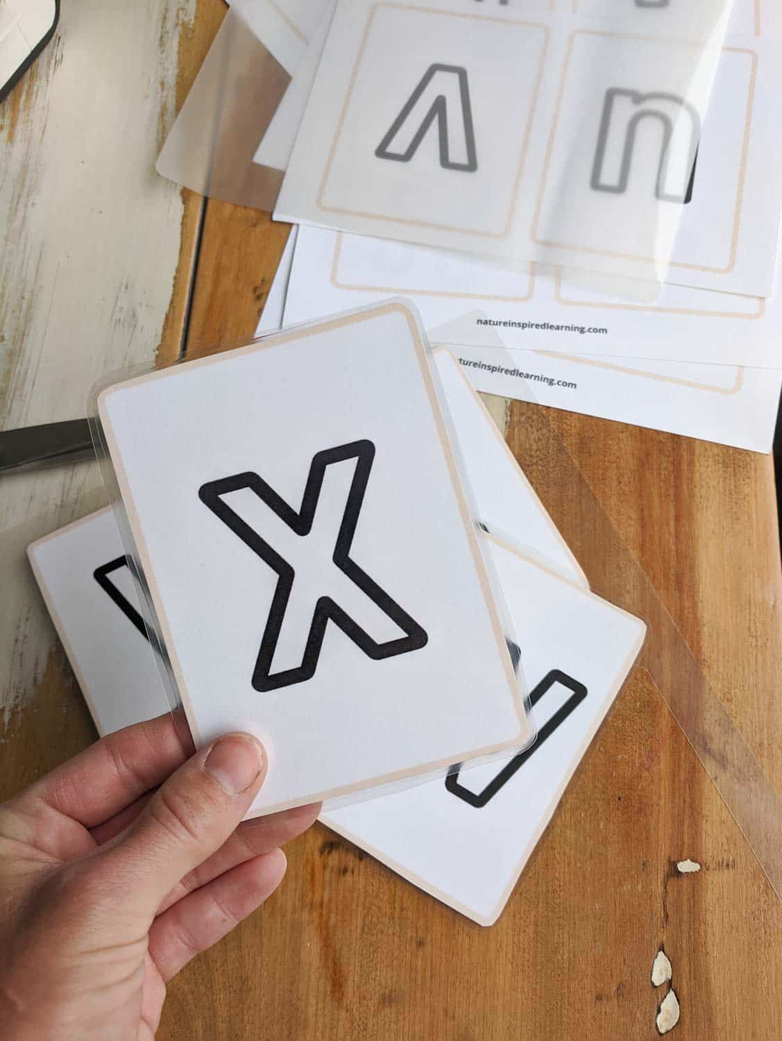 hand holding a laminated lowercase letter x flashcard over cut out cards on a wooden table. Plastic laminating sheet on top of a pile of printed out cards.
