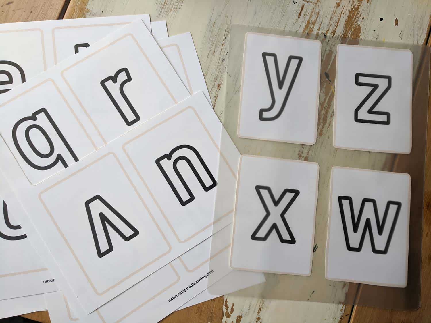 printed off flashcard set with lowercase letters in a pile on a wooden table to the right letters y, z, x, and w in a laminating sheet