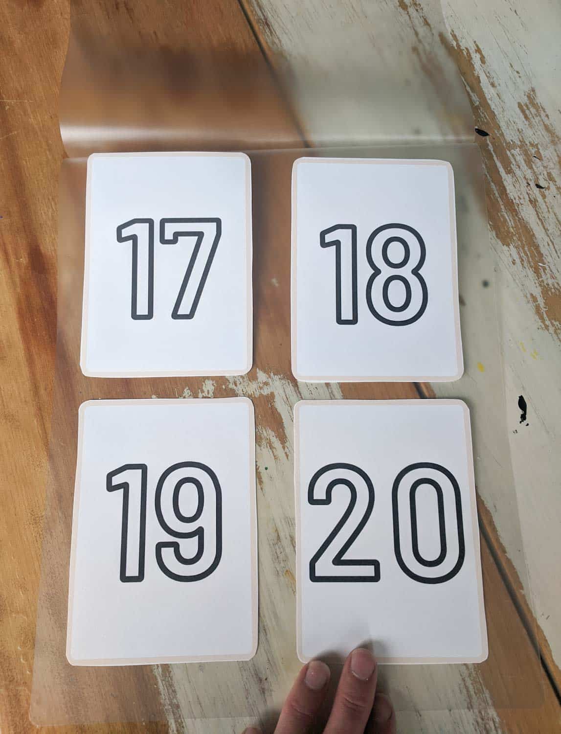 number cards 17, 18, 19, and 20 cut out and placed on a laminating sheet on a wooden table with white paint hand touching bottom right card