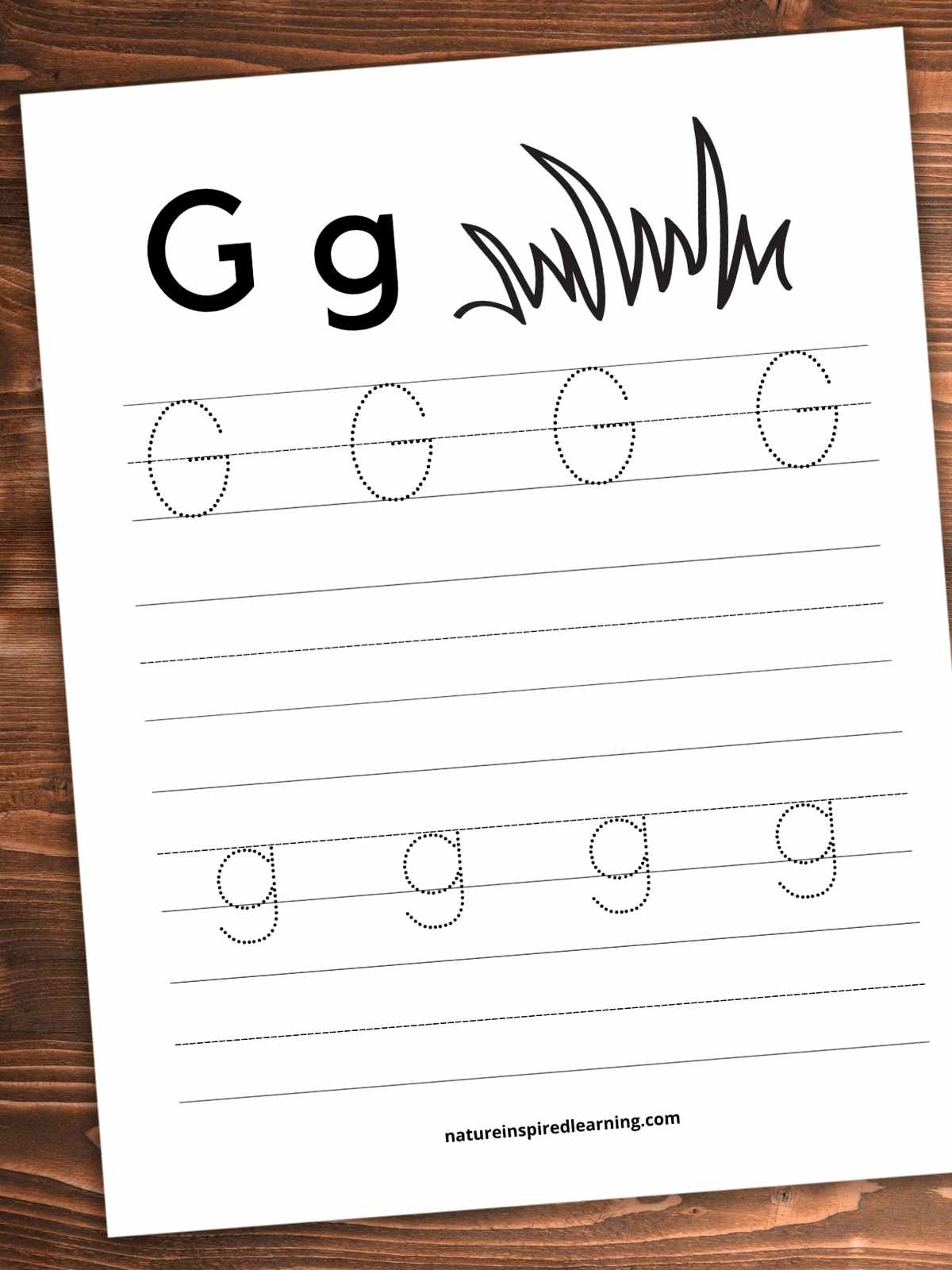 Worksheet with black and white blades of grass and bold G g across the top with a set of lines with dotted letter G's, a blank set of lines, a set of lines with dotted g's, and a blank set of lines across the bottom. Printable slanted on a wooden background