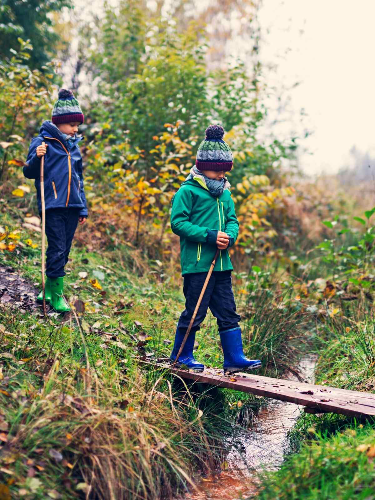 two children bundled up in jackets, rain boots, and winter hats walking outside in a natural fall landscape. Both children holding walking sticks one walking on a wooden board across a small stream